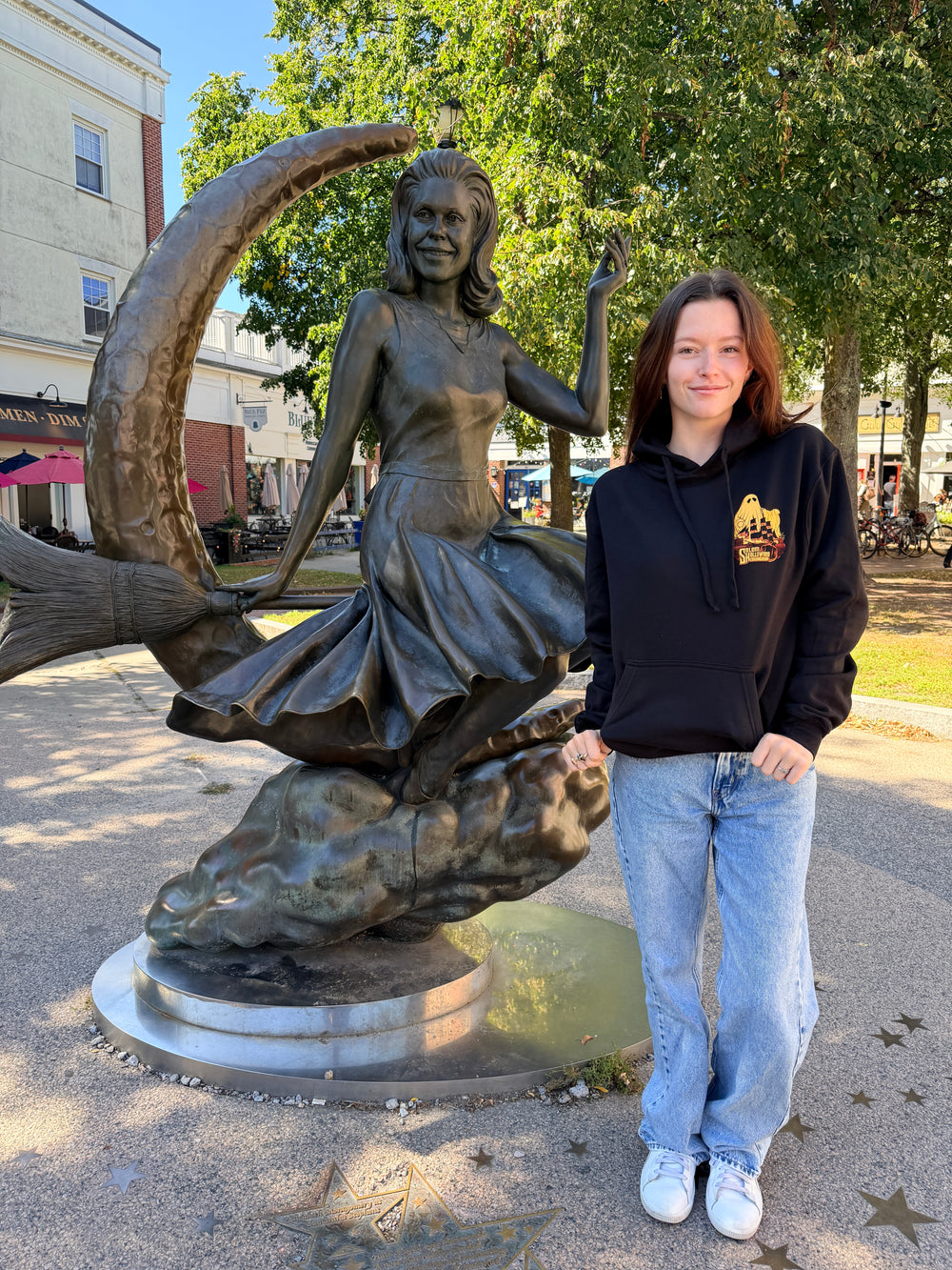 Front of the black 2026 Kaiju Ghost hooded sweatshirt, modeled beside the Bewitched statue in Salem.