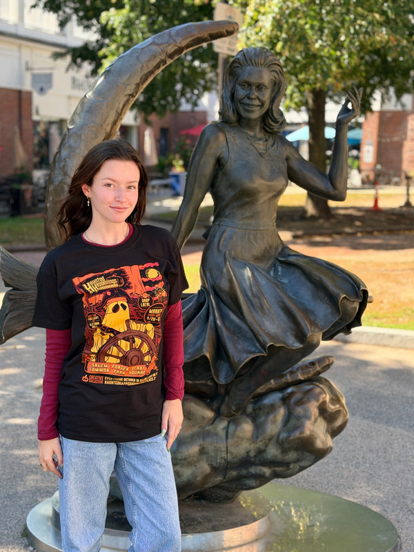 Model wearing the black 2026 Pirate Ghost T-shirt beside the Bewitched statue in Salem.
